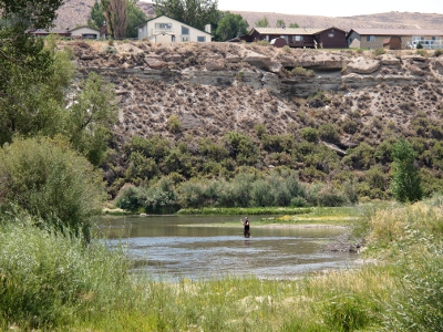 [A man stands in water reaching mid-thigh with one arm extended as he has just cast his pole. Behind him is the high, rocky bank with houses atop it. The water is surrounded by green vegetation.]